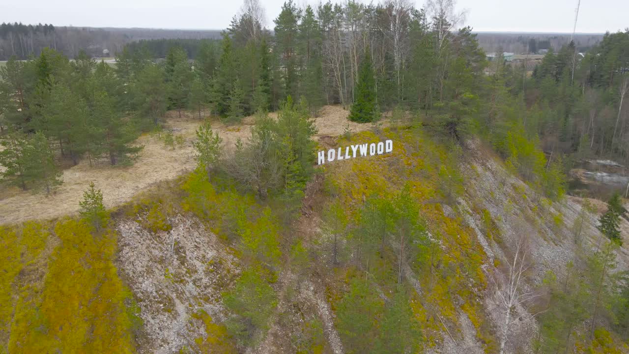 Aerial drone footage orbiting and circling around a white sign that Says Hollywood in Tartumaa Estonia called Hollywoodi mägi during spring time cloudy day. The sign is on a gravel mossy mountain.