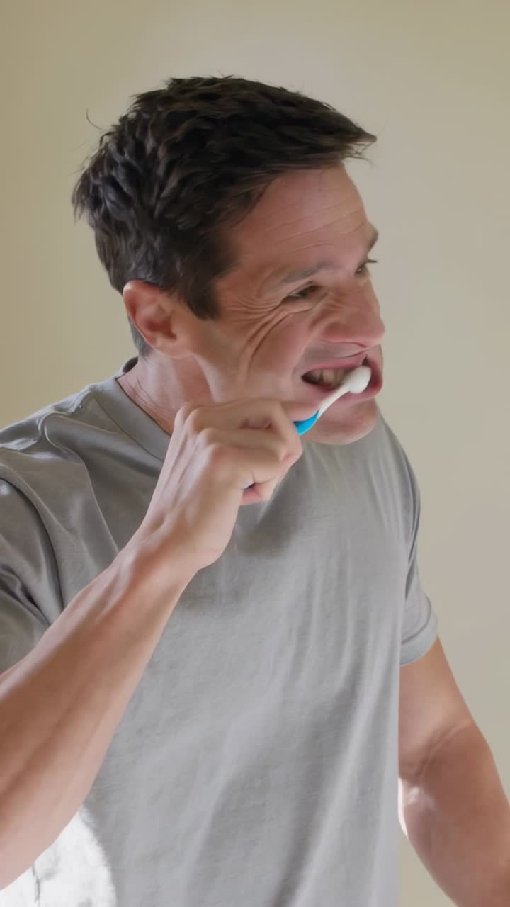 Vertical video: Grabbing blue brush, man in gray tee brushing teeth at bathroom sink, foam forming