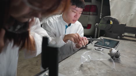 partial view of lady putting on face shield while observing microscope and young technician adjusts vernier caliper, taps on tablet in lab with carburetor on table and white car parked in background