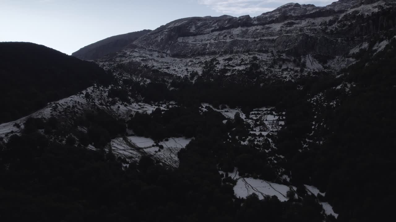 This drone video features a stunning reverse descriptive shot over the Hecho Valley in the Aragonese Pyrenees, Spain, revealing snowy mountains at sunset under a cloudy sky, creating a dramatic scene.