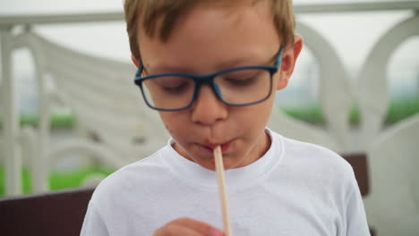 a young boy with glasses enjoys chocolate milk through a straw, with remnants of chocolate on his mouth, a partial view of another child is visible in the background