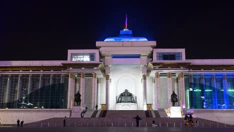 during a calm nighttime scene, people explore sukhbaatar square in ulaanbaatar, where a bronze statue of genghis khan, the mongolian emperor, stands before the government palace with a flag above.
