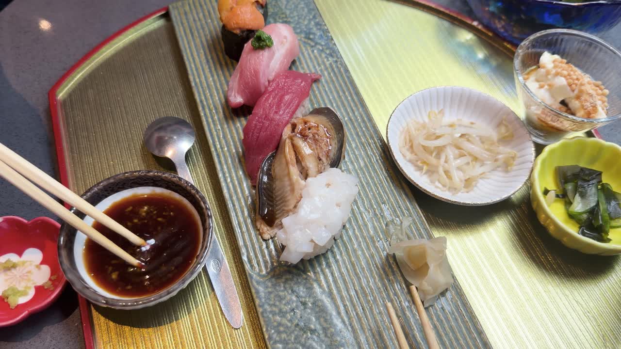 Overhead view of assorted nigiri sushi platter as chopsticks dip into soy-based sauce bowl