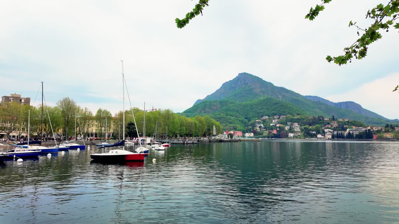 Inquadratura grandangolare di Lecco, Italia, che mostra barche a vela ormeggiate sul tranquillo Lago di Como, montagne verdi e lussureggianti, città affascinante, luce primaverile limpida, lungolago tranquillo, nessuna persona visibile.