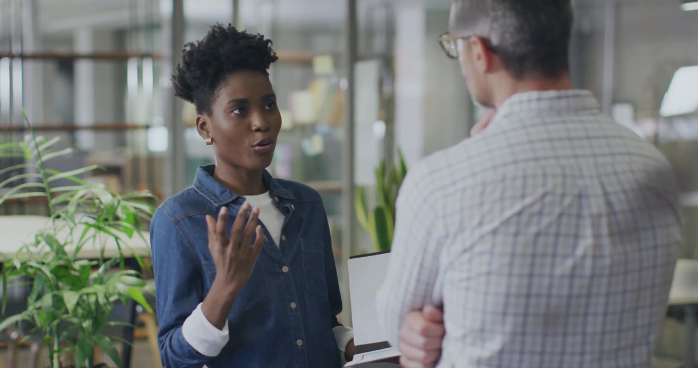 Vertical video: Facing man, woman in denim pointing at laptop explaining in office, glass partition