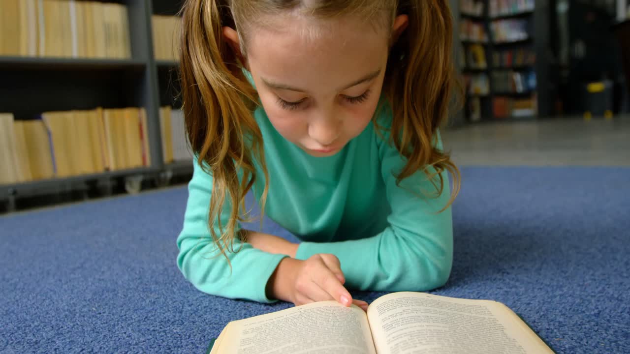 Vista frontale di una scolaretta caucasica attenta che legge un libro in biblioteca a scuola 4k