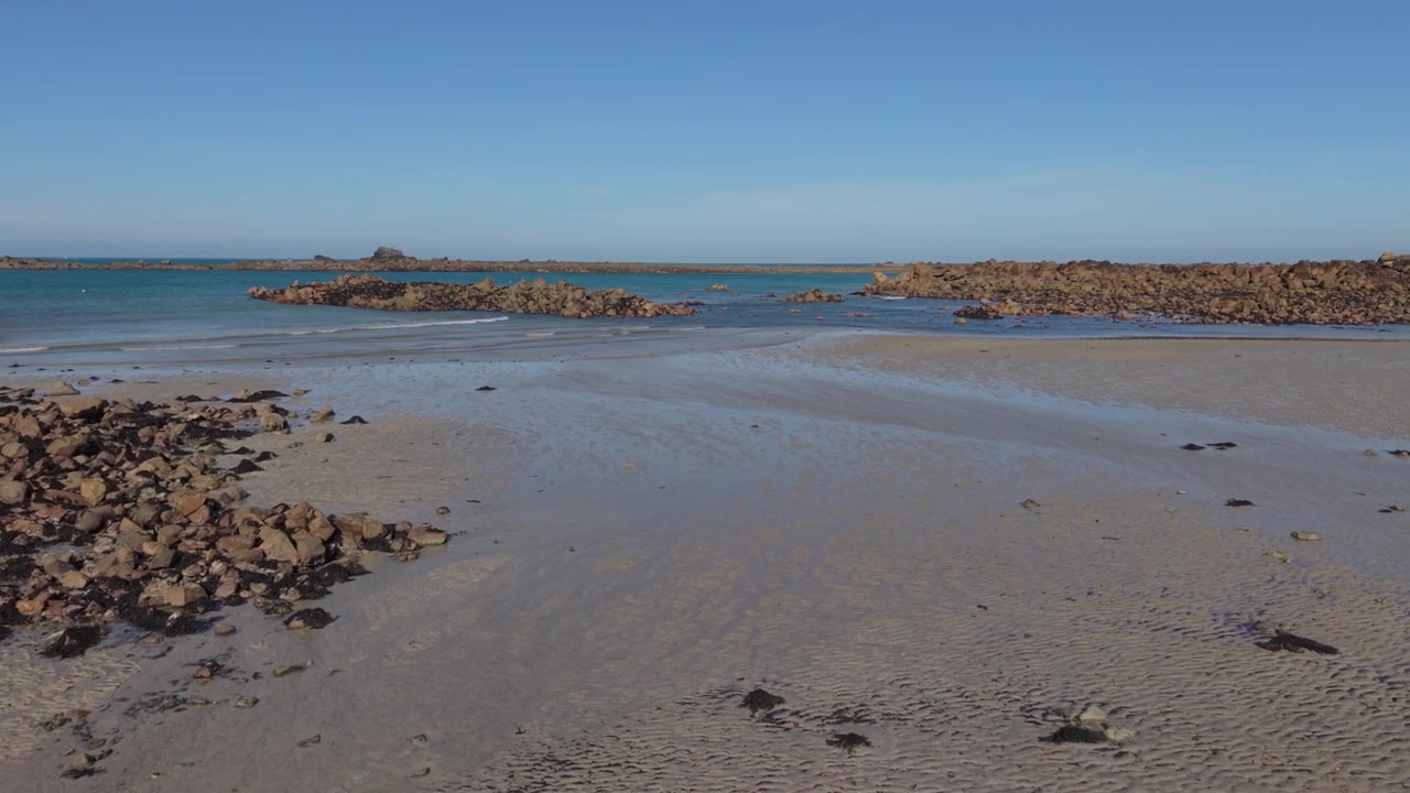 Guernsey, Normanské ostrovy. Pomalé záběry dronu vpřed za odlivu s jemnými vlnami přicházejícími na břeh mezi skalními výchozy přes písčité pobřeží drsné skalnaté pláže v pozdním odpoledním slunci.