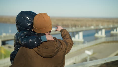 a father carries his son while standing by an iron railing, showing him something across the bridge, with blur view of cars passing by on the bridge