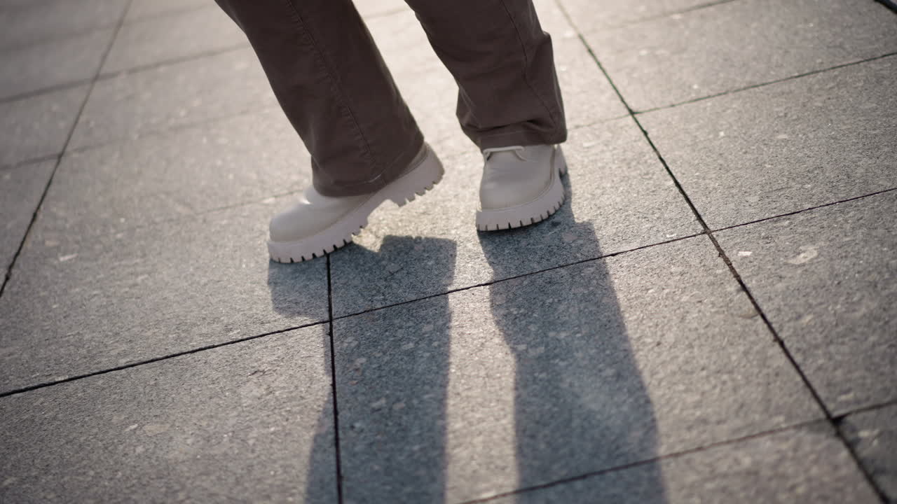Closeup of dancer legs in beige corduroy pants and chunky white platform shoes moving rhythmically across sunlit tiled plaza with snow piles in background, casting long shadows on cold winter ground