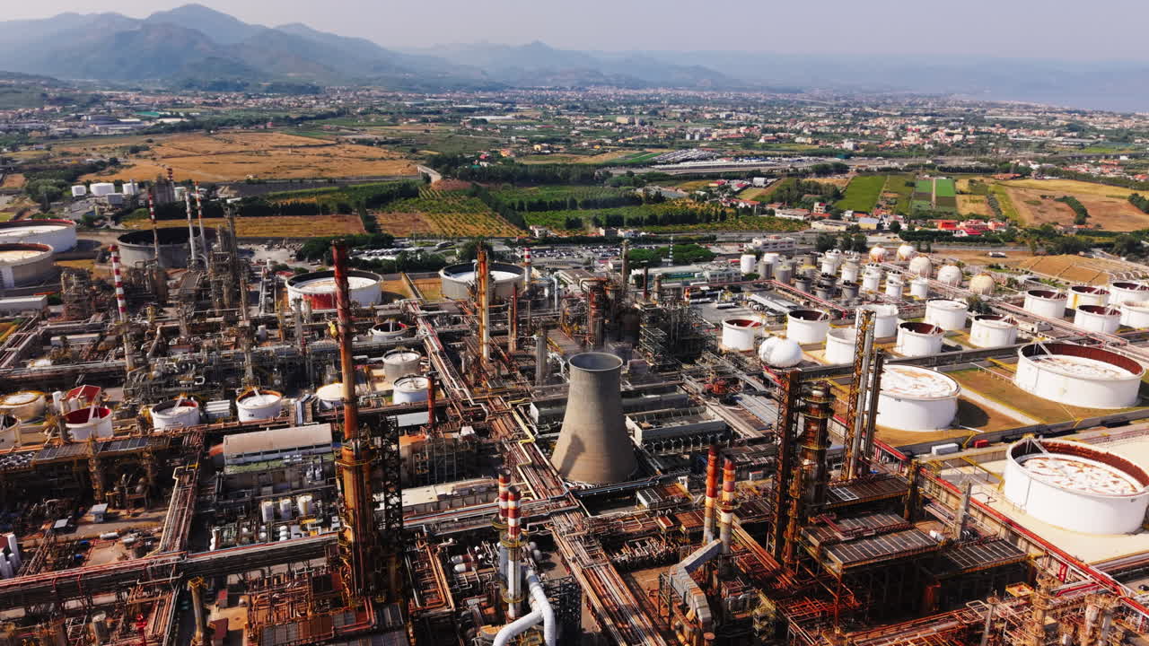 Drone descends and tilts up over an oil refinery in Sicily, showing tanks, chimneys, and distant fields