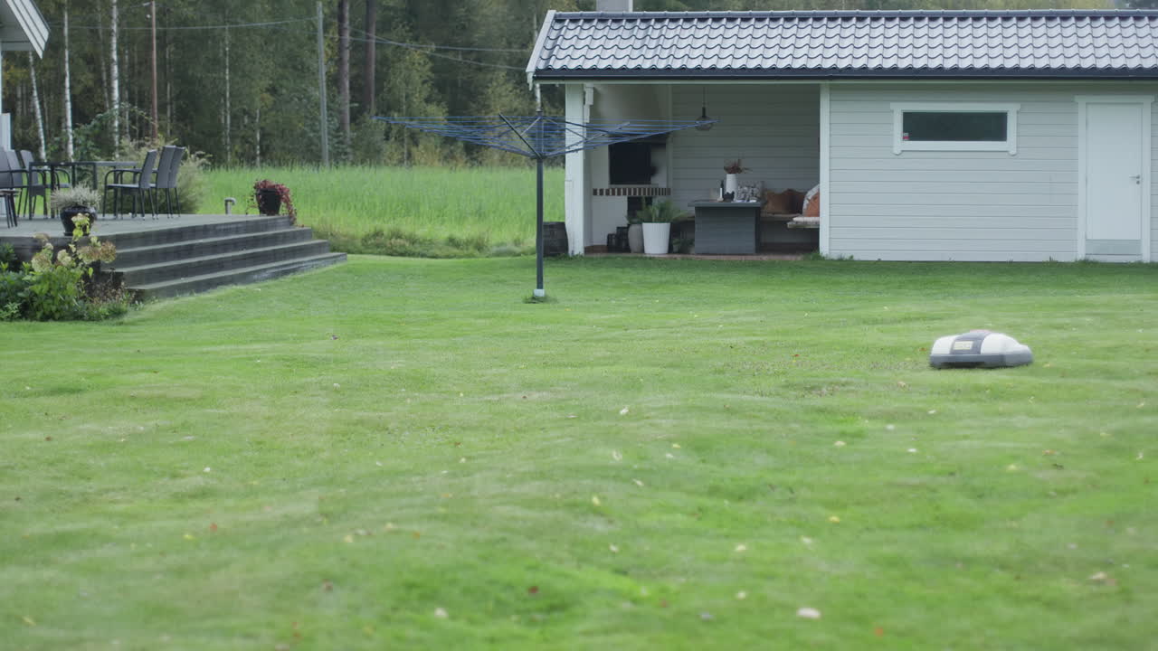 A robotic lawn mower on green grass near a modern patio and garden seating area with a shed.