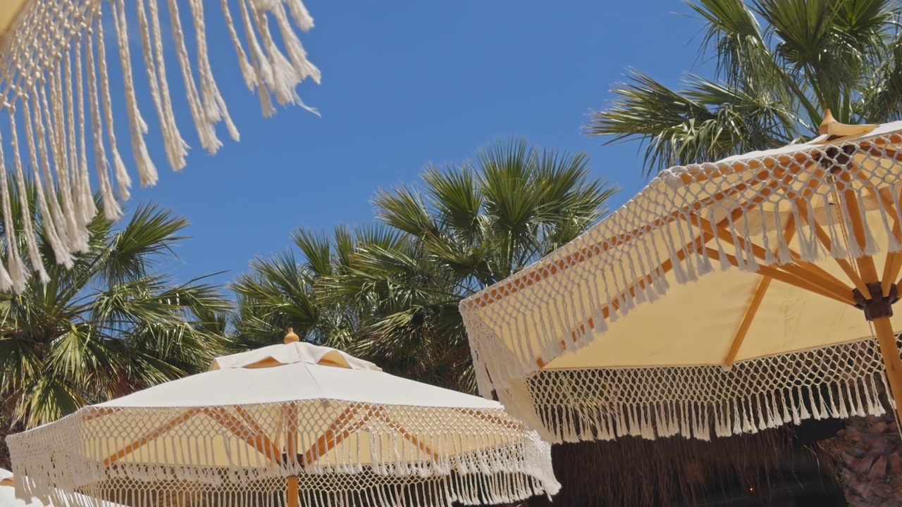 Inquadratura media statica di ombrelloni boho chic con frange che ondeggiano dolcemente con palme tropicali e un cielo azzurro chiaro sullo sfondo in un resort di lusso sulla spiaggia.