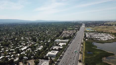 Aerial-view-of-suburbs-dense-trees-rooftop-houses-pine-trees-cars-freeway-101-marsh-on-left-pivot-right