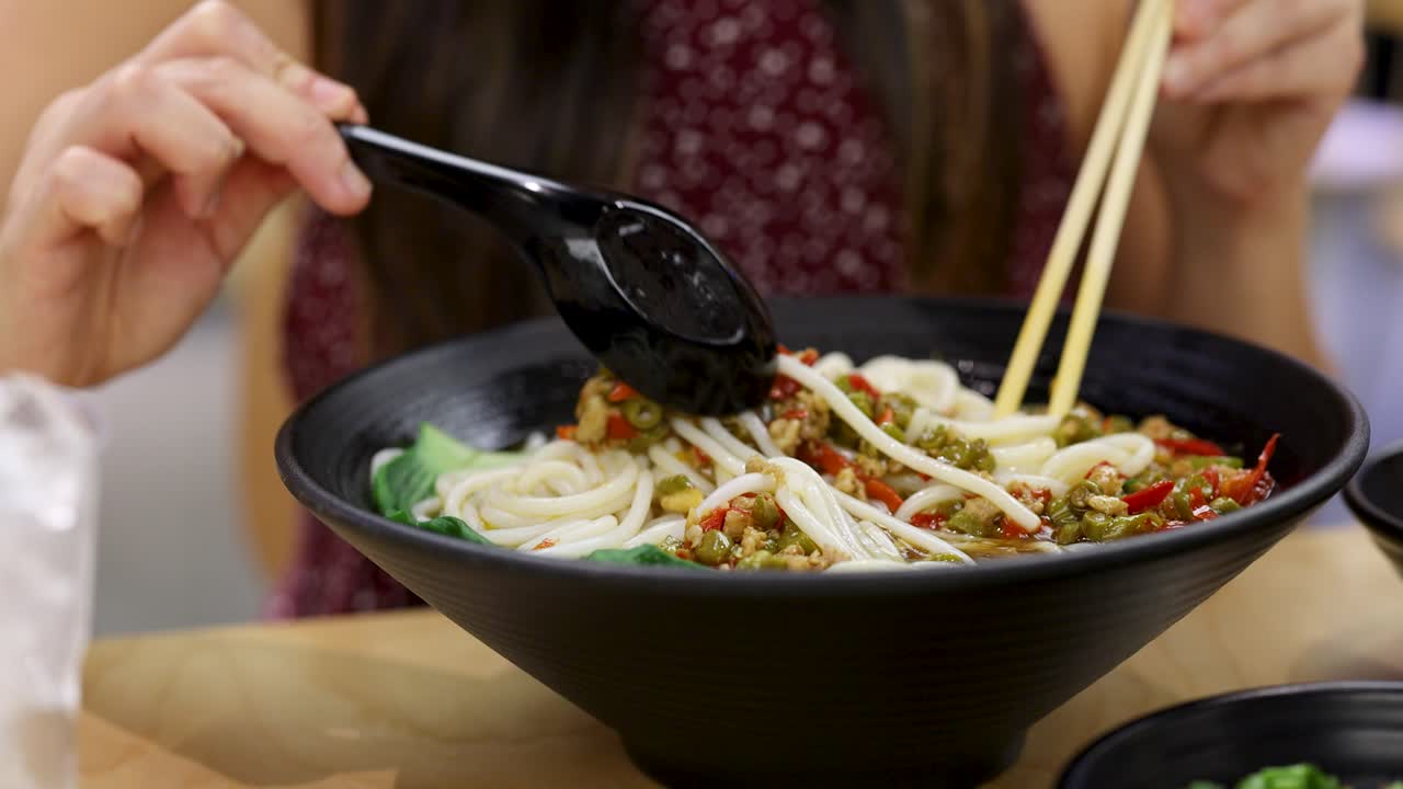 Person stirs spicy Chinese noodle soup in black bowl, bright indoor lighting, close-up view