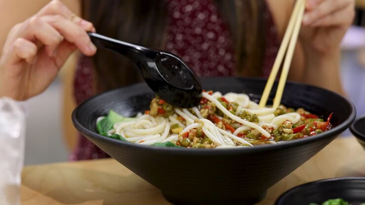 Una persona prepara una zuppa di noodle cinesi piccante in una ciotola nera, illuminazione interna intensa, vista ravvicinata