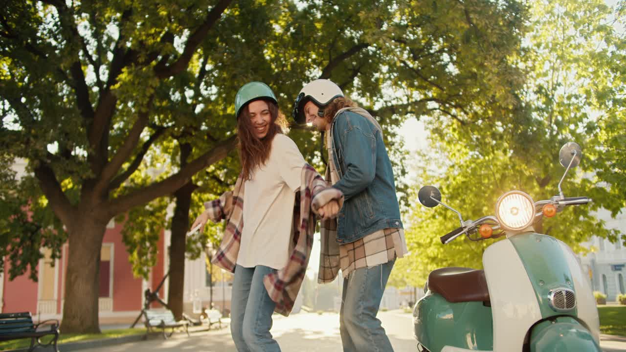 Happy girl in a checkered shirt and a motorcycle helmet in denim pants dances with her boyfriend in a denim jacket near her green moped in a sunny summer park in the city