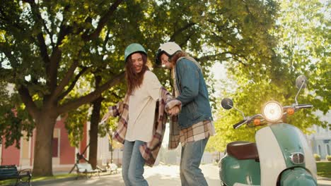 Happy-girl-in-a-checkered-shirt-and-a-motorcycle-helmet-in-denim-pants-dances-with-her-boyfriend-in-a-denim-jacket-near-her-green-moped-in-a-sunny-summer-park-in-the-city