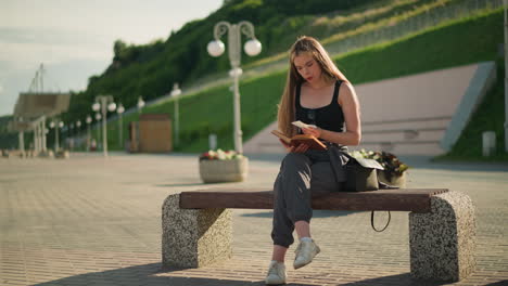 woman seated outdoors on a bench with legs crossed, flipping a page from her book and observing a piece of paper thoughtfully, the background features lamp poles and a person seated in the distance