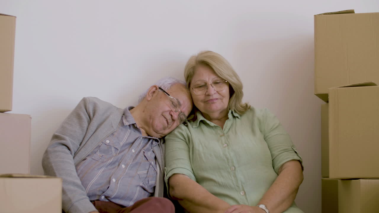 Front view of tired and cheerful elderly wife and husband sitting on floor with carton boxes after moving