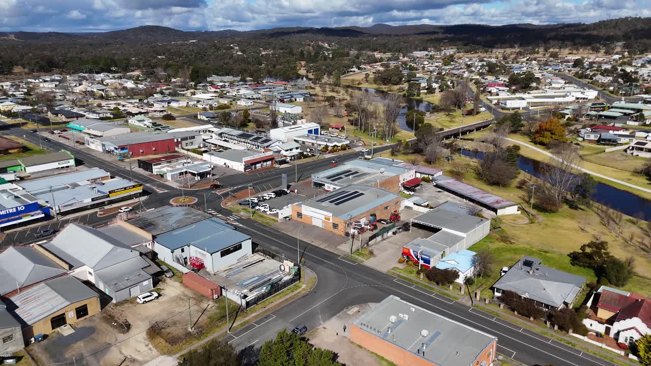 Imagens de drone deslizam sobre uma tranquila cidade australiana, revelando casas residenciais, carros estacionados e edifícios comerciais sob luz do dia intensa com nuvens dispersas.