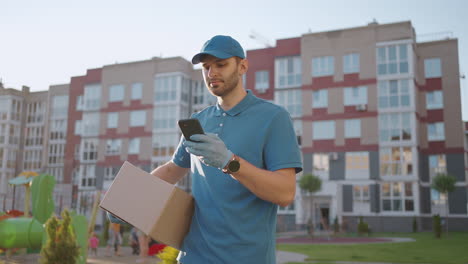 El-Cartero-Con-Gafas-Lleva-El-Paquete-Y-Mira-La-Dirección-De-Entrega-A-Través-Del-Teléfono-Móvil.-Buscar-La-Dirección-Del-Cliente-De-Entrega.-Repartidor-Con-Gorra-Y-Caja