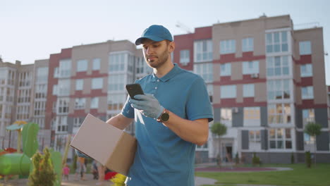 el cartero con gafas lleva el paquete y mira la dirección de entrega a través de un teléfono móvil. busque la dirección del cliente de entrega. el tipo de entrega con una tapa y una caja