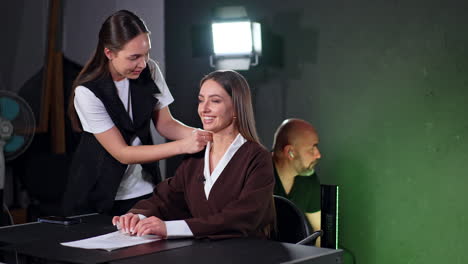 beautiful smiling lady sitting at desk is prepared for video shooting. assisting girl fixing reporter's clothes. male photographer sits at backdrop.