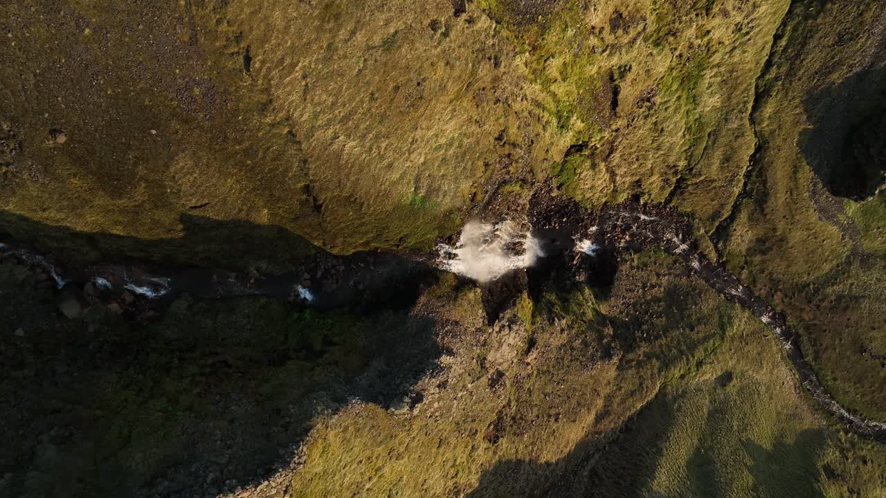 Aerial shot of a small waterfall flowing through rugged terrain in northern Iceland, surrounded by moss-covered land and untouched wilderness.