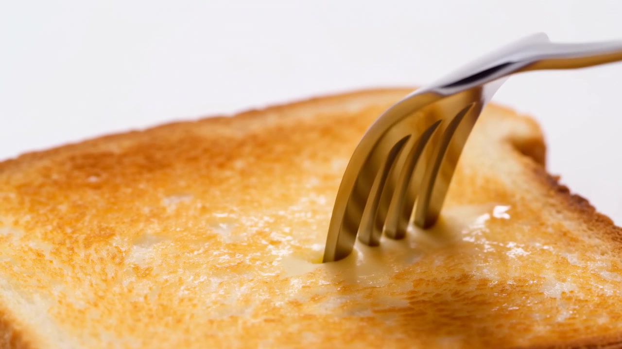 Motion of fork spreading melted butter across golden toasted bread, with visible dragging, soft pressing, glossy streaks, and shallow crumb marks in bright appetizing breakfast closeup