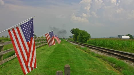 a view of a line of gently waving american flag on a fence by farmlands as a steam passenger train blowing smoke approaches in late afternoon