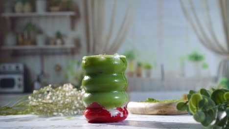 a hand with natural nails and silver rings gently reaches for a colorful iced matcha strawberry latte in a sculpted glass, set on a rustic table with green plants and matcha powder nearby.