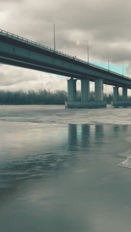 grey bridge over wide river with ice against dense forest under blue sky with clouds on winter day aerial view