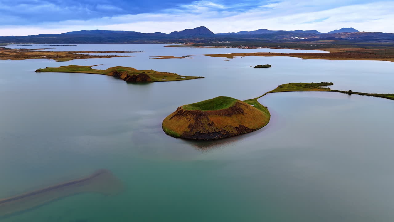 Small island with volcano crater on the water surface. Silhouettes of mountains at the waterfront at backdrop. Scenery of Iceland from top.