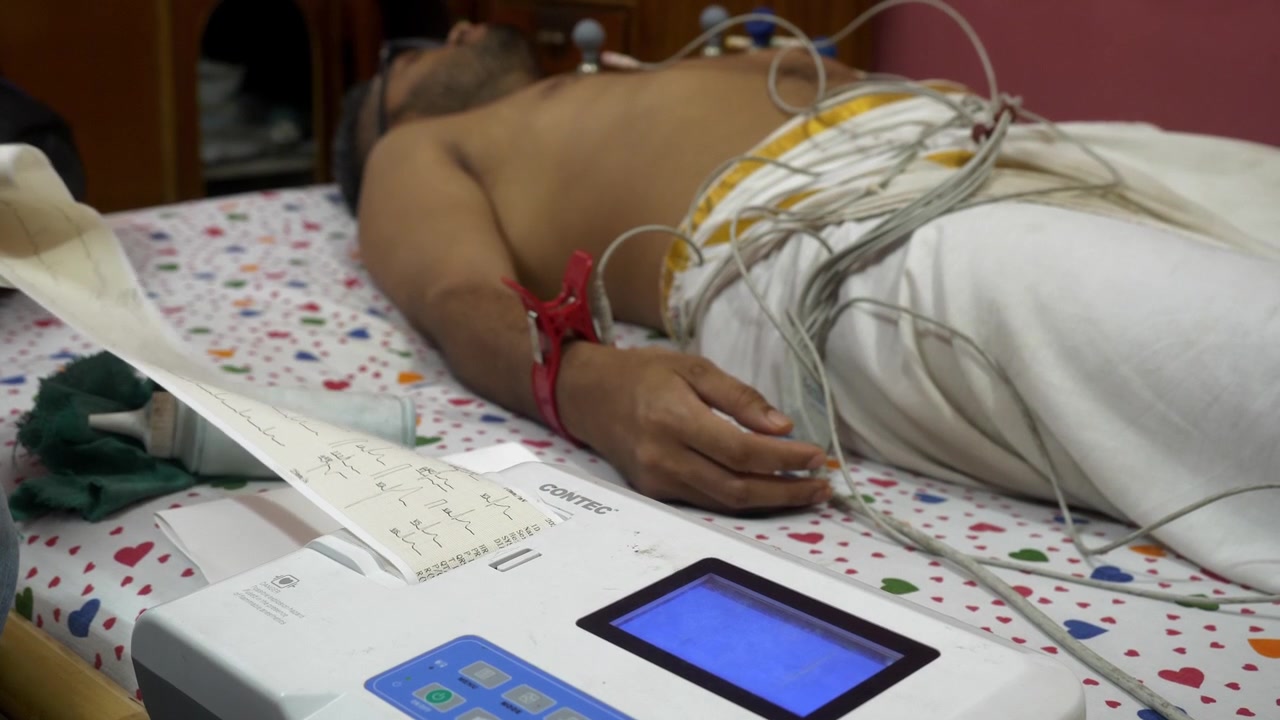 Elettrodi della macchina ECG attaccati al petto e alle braccia del paziente per registrare i segnali elettrici del cuore in un ambiente domestico, con carta millimetrata del referto ECG.