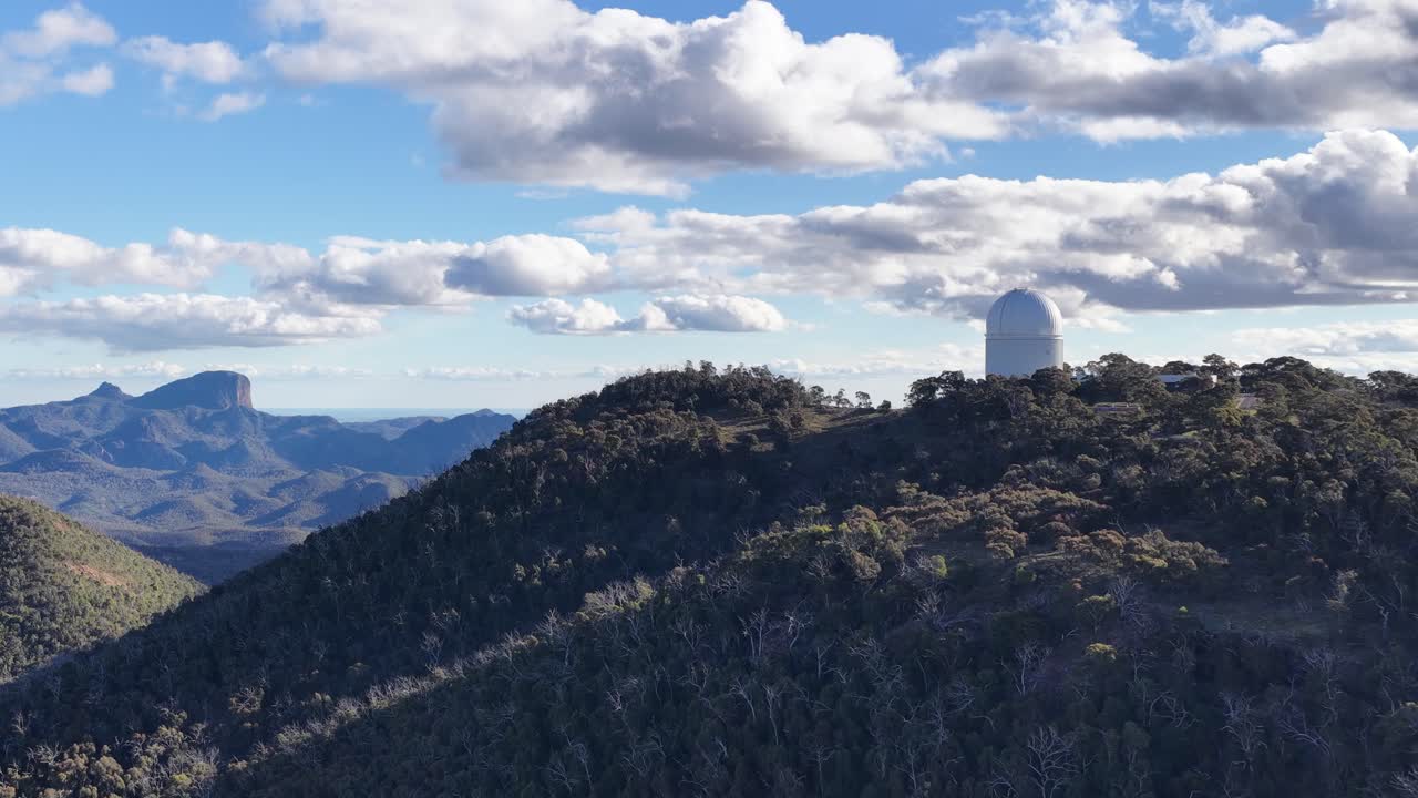Drone ascends above forested hills, revealing Siding Spring Observatory atop a ridge under bright daylight with scattered clouds and expansive mountain views