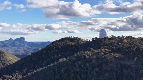 drone ascends above forested hills, revealing siding spring observatory atop a ridge under bright daylight with scattered clouds and expansive mountain views