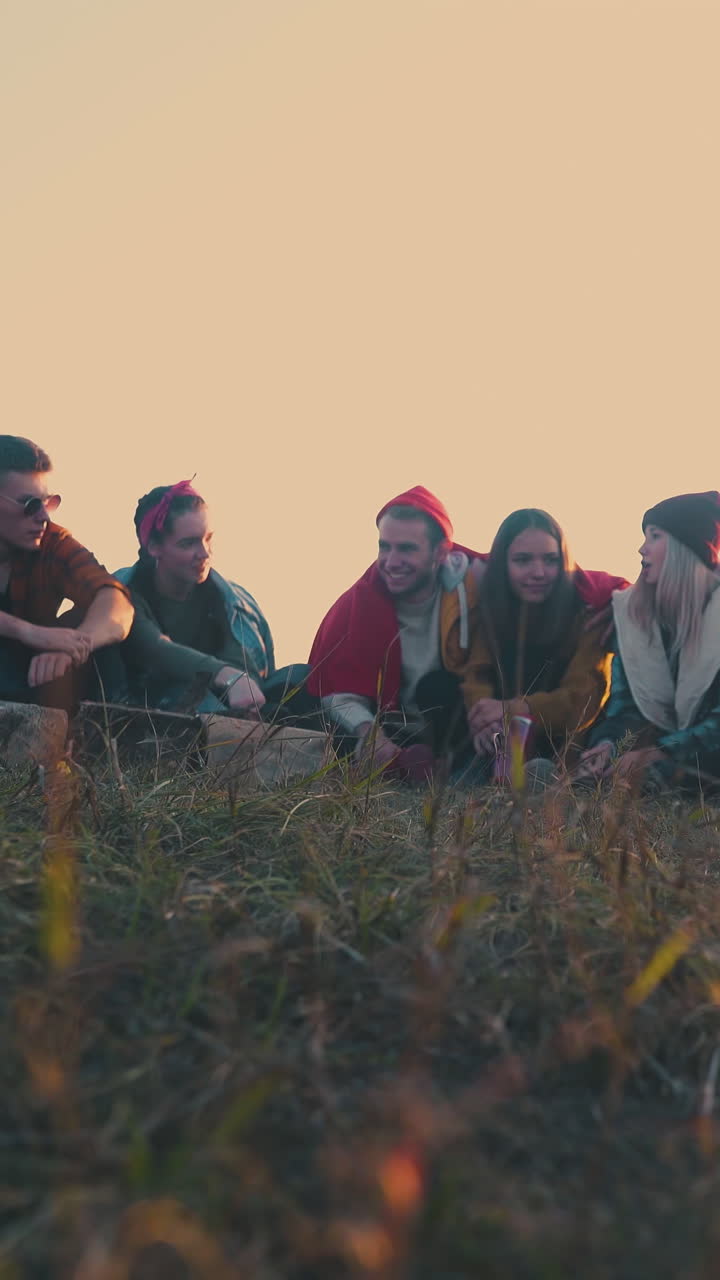 happy young tourists talk sitting at burning bonfire and tent on meadow at sunset in autumn evening low angle shot