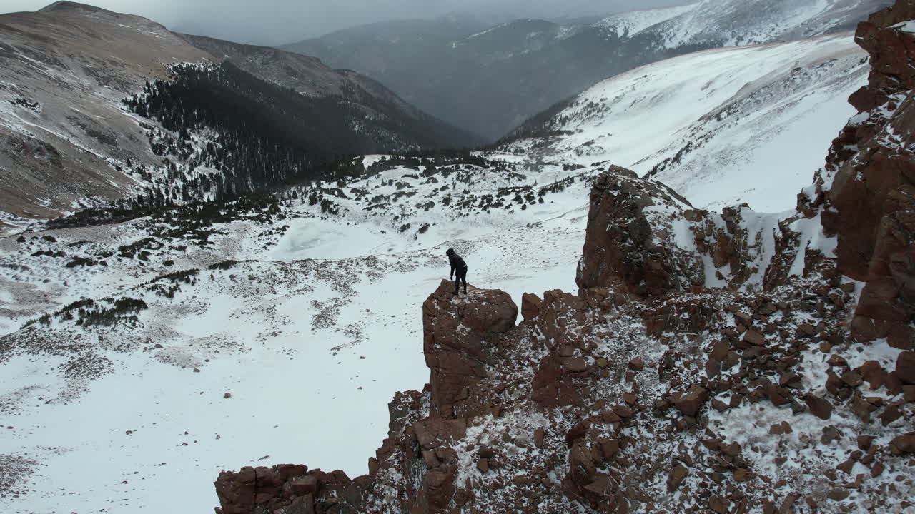 추운 겨울날 눈 덮힌 계곡 위의 바위 꼭대기에 서있는 남자 아드레날린 마약 중독자의 공중 전망, 드론 샷