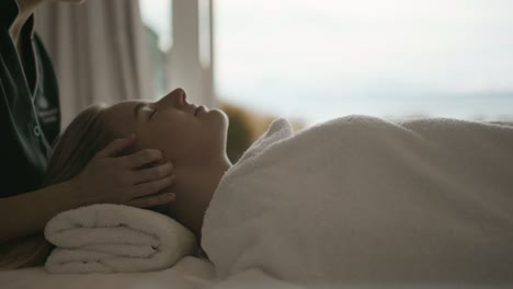 Mujer-Rubia-Recibiendo-Un-Masaje-Facial-Frente-A-Una-Gran-Ventana