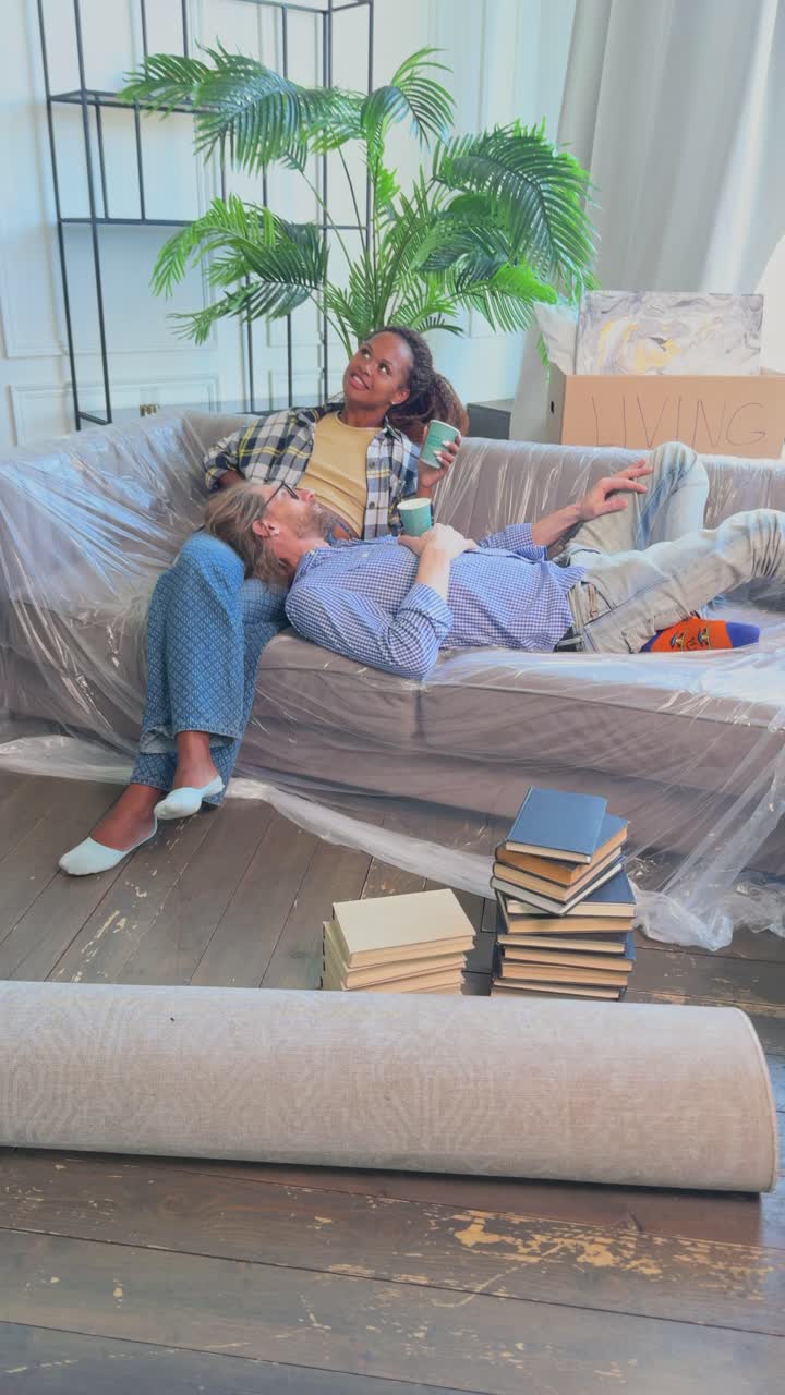 una pareja joven y diversa, mujer y hombre, se relajan en el sofá después de mudarse y beben café.