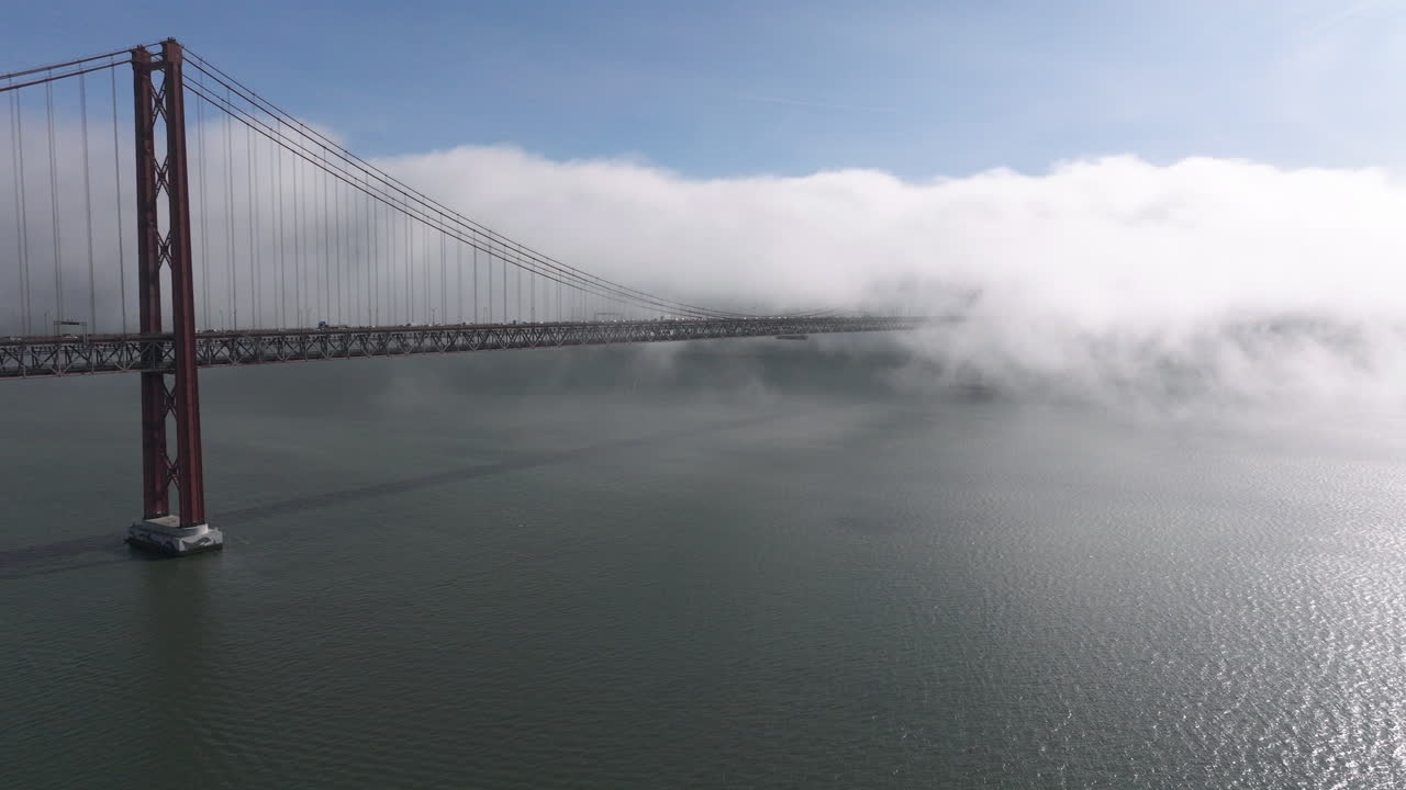 Aerial drone shot towards the 25th April Bridge and waterfront in Lisbon, Portugal, Europe. Sunny and bright with clouds, fog, mist covering the bridge. Shot in ProRes 422 HQ