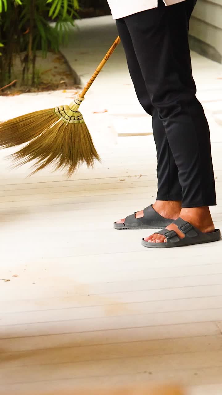 A person sweeps a wooden deck with a broom, surrounded by wooden furniture and greenery, under natural daylight