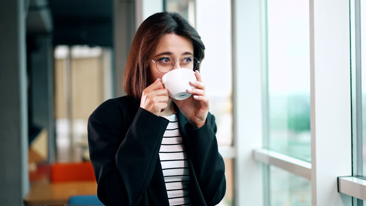Thoughtful young woman wearing glasses stands at the panoramic window. Lady drinks coffee during the work break.