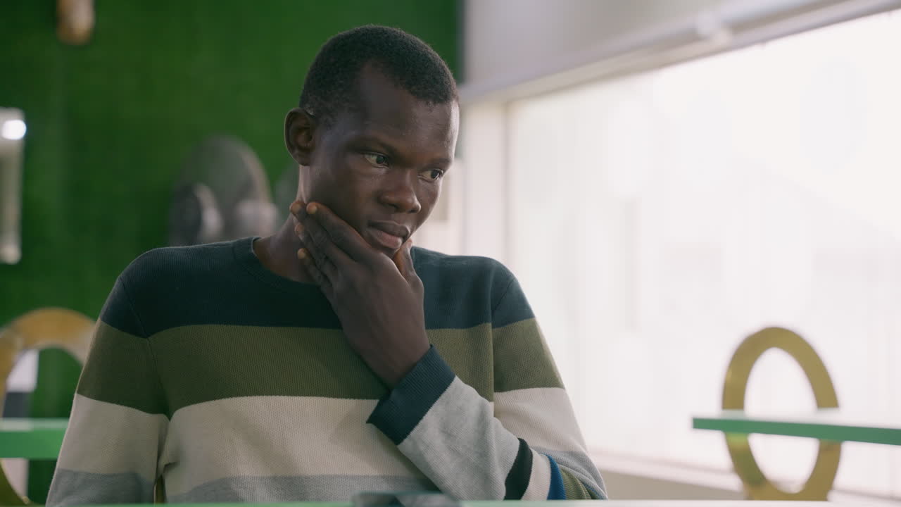 Young black man thoughtful in cafe, touching chin and staring past window with green backdrop striped sweater, quiet table, subtle tension between hope and doubt as he considers next move