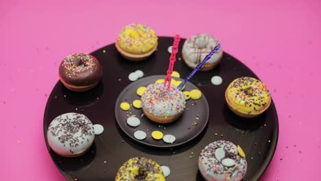a bunch of american donuts with black, yellow and blue chocolate and choco and multicolored crumb toppings rotates on a pink and black background in a curcle. shot from above