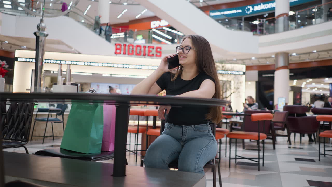 쇼핑몰에서 테이블에 앉아있는 젊은 여성, 행복하게 그녀의 에 쇼핑 가방과 함께 전화를 하고, 다른 사람들은 주변에 앉았고, 배경에 biggies 표지판이 보이며, 활기찬 분위기를 만니다.