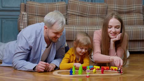 Family-of-mother,-father-with-daughter-child-girl-riding-toy-train-on-wooden-railway-at-home-room