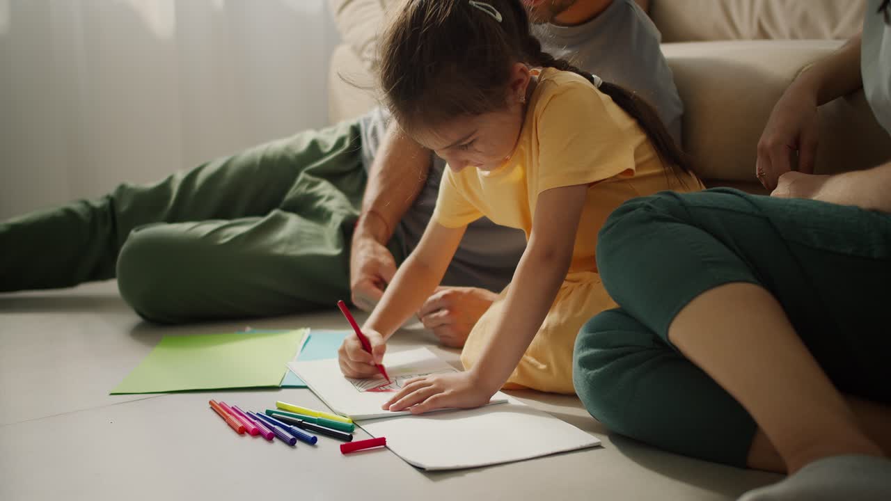 fotografía en primer plano de una pequeña chica morena feliz en un vestido amarillo sentada en el suelo cerca de sus padres y dibujando en papel usando bolígrafos de punta de fieltro multicolores en un apartamento moderno