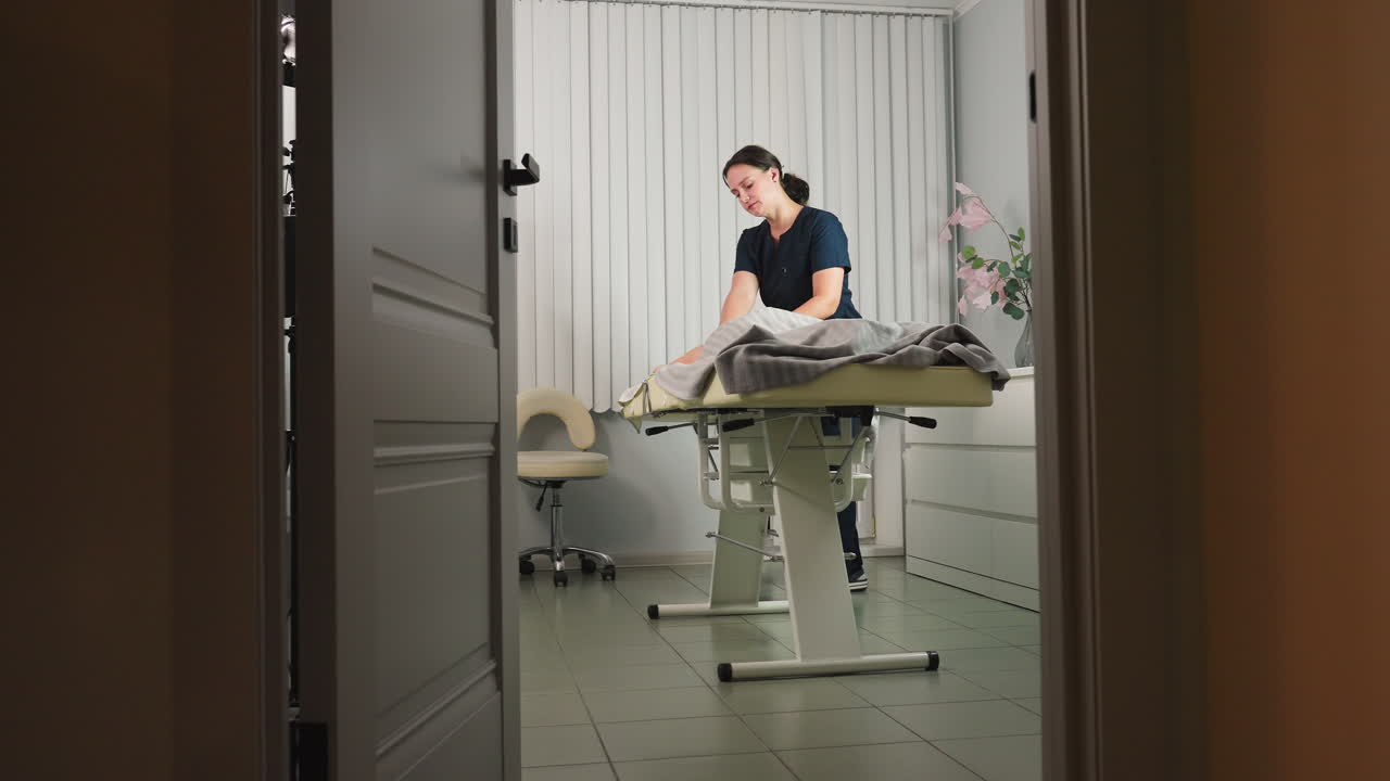 Door view of modern spa room with skincare specialist in uniform massaging back of relaxed client lying face down on adjustable massage bed under soft lighting during a spa session