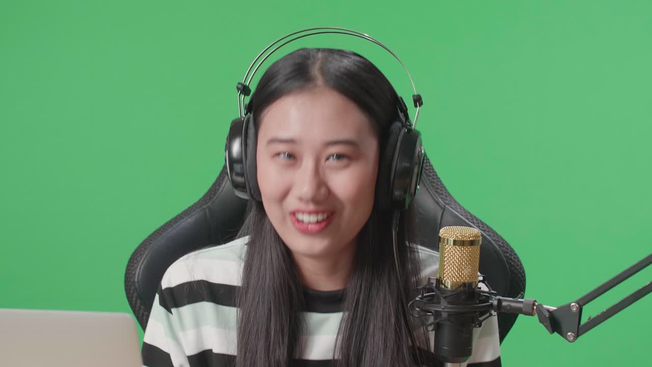 Close Up Of Asian Woman Gamer With Headphone And Computer Waving Hand Then Raising Index Finger While Playing Game On Mobile Phone On Green Screen